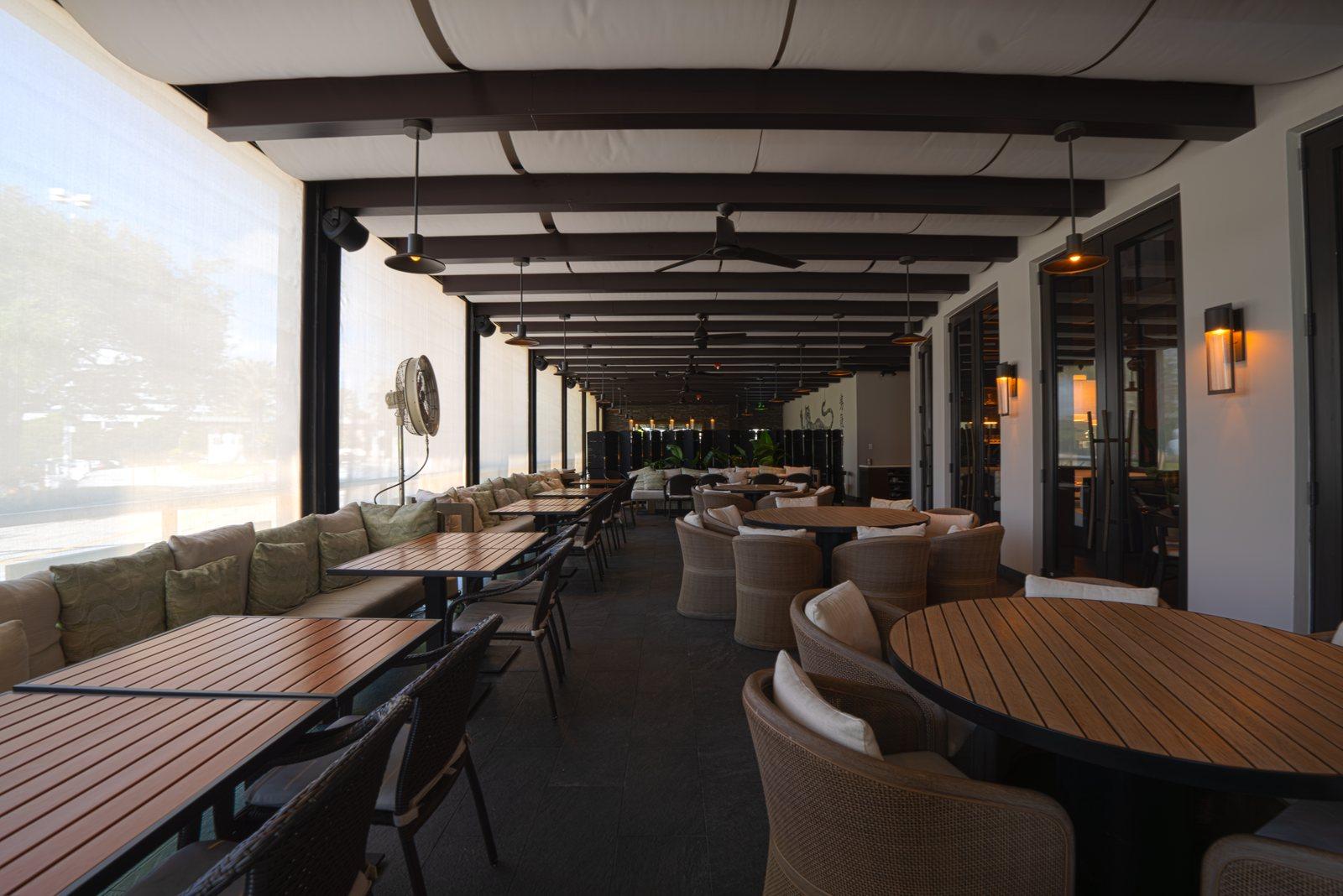 Covered restaurant patio with sun shades along the open side and teak tables