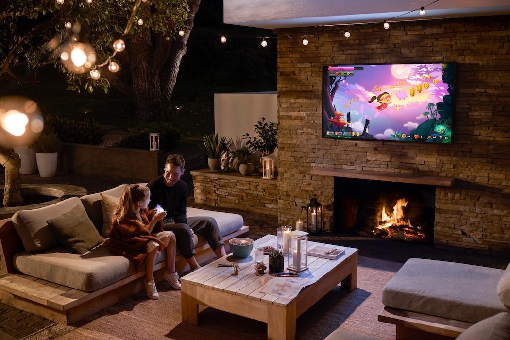 Covered outdoor patio at night with a wall-mounted TV above a stone fireplace and string lights